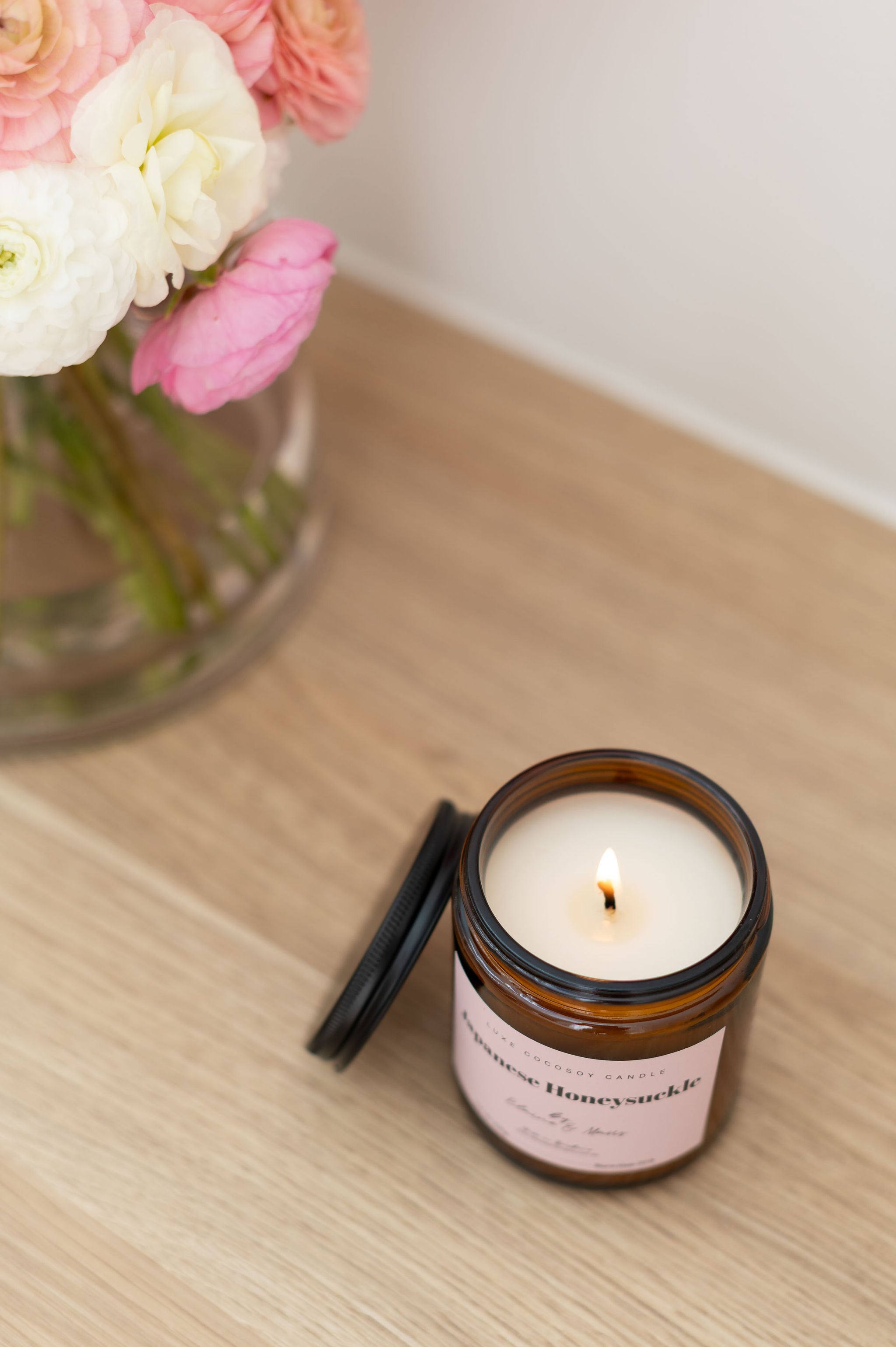 Candle in a glass jar with a label next to a vase of flowers on a wooden surface
