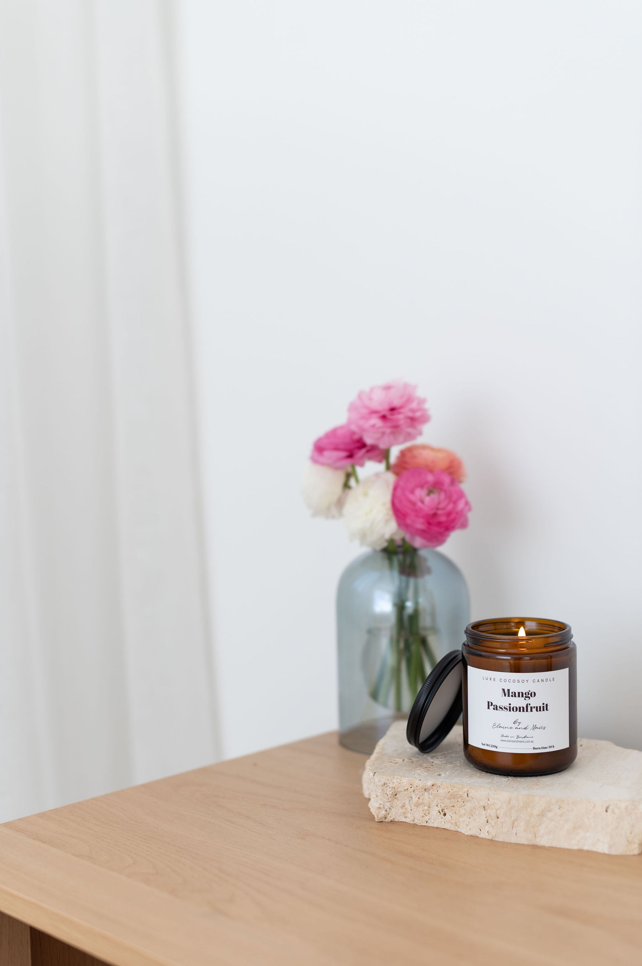 Soy Candle labeled 'Mango Passionfruit' on a stone coaster with a vase of flowers in the background.